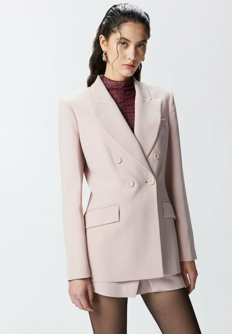 Pudrasto ružičasta dvoredna blejzer s izrezom na reverima, dva prednja džepa i odgovarajuće šorts hlače. Noseno preko maroon bluze s točkicama.