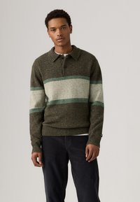 Strikkegenser i mørkegrønn, med horisontale striper i lysere grønn og beige. Krage med knapper og ribbestrikkede mansjetter med logo-detalj.