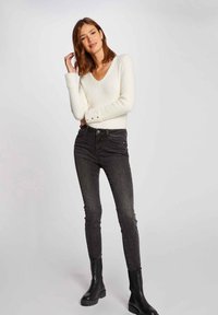 Weißer V-Ausschnitt-Pullover aus strukturiertem Stoff mit Knopfdetails an den Ärmeln, kombiniert mit hochgeschnittenen schwarzen Jeans und schwarzen Stiefeletten.