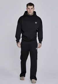 Felpa con cappuccio nera e pantaloni cargo abbinati in tessuto morbido. Caratterizzati da una tasca frontale, logo sul petto e tasche laterali sui pantaloni.