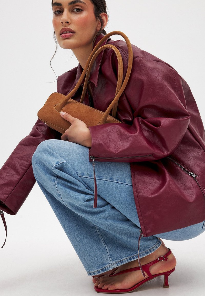 Burgundy faux leather jacket, blue wide-leg jeans, and red strappy heels. Brown suede handbag held against the jacket. Smooth texture.
