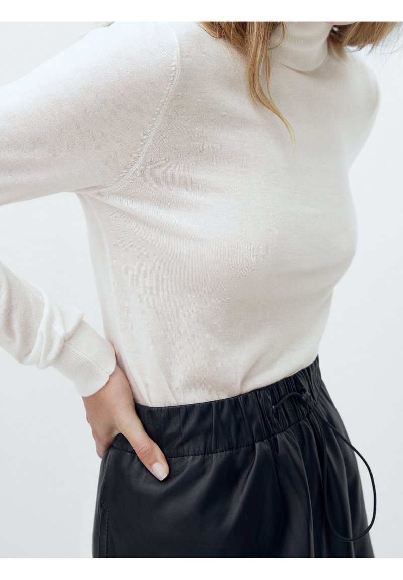 Kobieta w jasnobeżowym swetrze z golfem i czarnych skórzanych spodniach z elastycznym pasem, ręka oparta na biodrze, obcięta na ramionach.