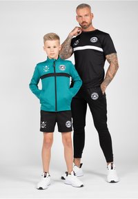 A boy in a teal zip jacket with black accents and logo, paired with black shorts. An adult in a black and white shirt with matching pants stands beside.