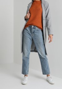 Un manteau en laine gris par-dessus un pull en tricot orange, un jean bleu droit et des baskets blanches. Le manteau a un col cranté et une fermeture à boutons.