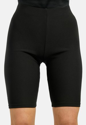 Pantalones cortos de ciclismo ajustados de color negro que cubren hasta la mitad del muslo, llevados en cintura y caderas sobre un fondo liso.