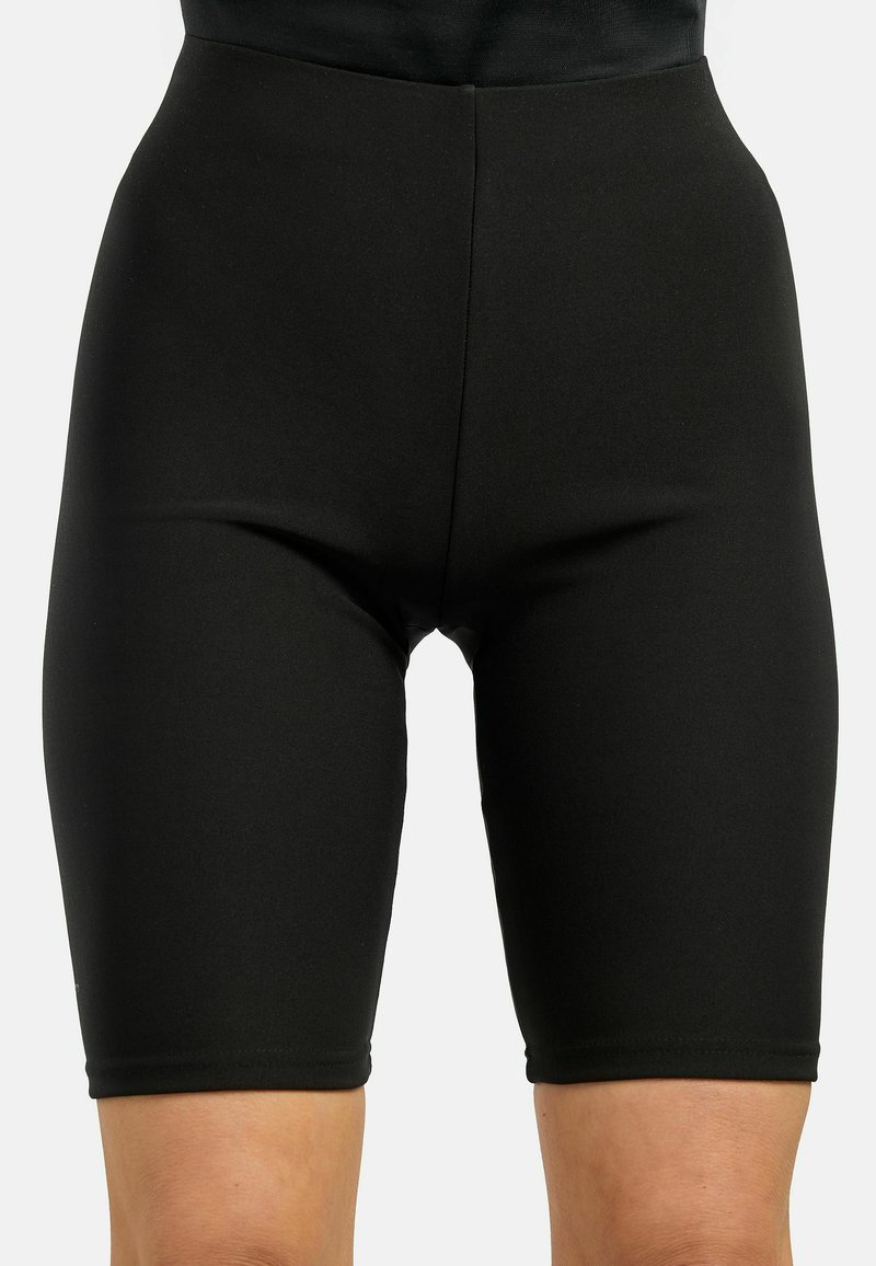 Cuissard de cyclisme noir moulant couvrant le milieu des cuisses, porté à la taille et aux hanches sur un fond uni.