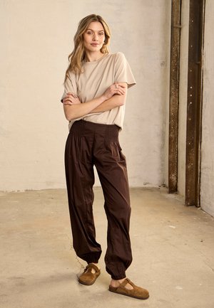 Femeie purtând un top bej cu mânecă scurtă, pantaloni maro închis cu talie elastică și pantofi slip-on bej, stând cu brațele încrucișate într-un spațiu interior neutru.