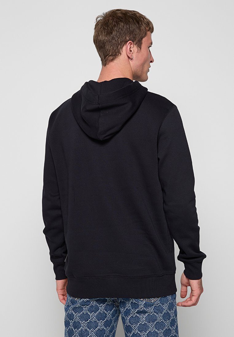 Mann mit dem Rücken zur Kamera, der einen schwarzen Kapuzenpullover und blaue gemusterte Jeans trägt, vor einem hellen einfarbigen Hintergrund.