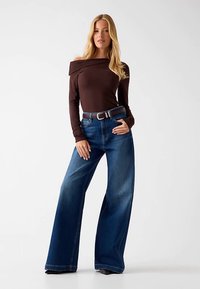 Haut à manches longues marron décolleté en bardot, associé à un jean en denim bleu foncé à taille haute, jambes larges, et une ceinture marron.
