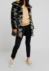 Abrigo de piel sintética en gris con patrón de camuflaje negro, con capucha, llevado sobre una sudadera beige y pantalones negros. Zapatillas color tostado completan el conjunto.