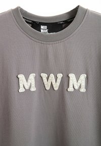 Camiseta de algodón gris con tres letras blancas texturizadas "M", "W" y "M" en una fuente decorativa, ubicadas en la parte frontal.
