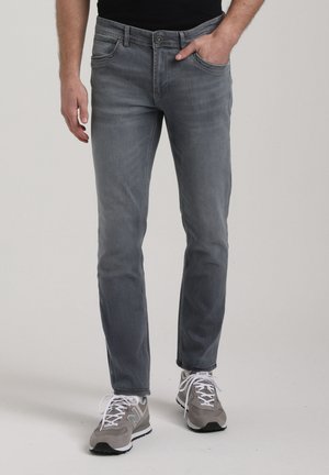 Mann trägt schmal geschnittene graue Jeans und graue Sneaker, mit einer Hand in der Tasche, steht vor einem schlichten hellen Hintergrund.