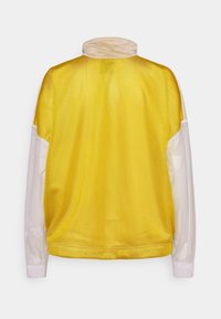 Gul meshjacka med hög krage, vita ärmar, elastiska muddar och avslappnad passform. Lätt material med en andningsbar design.