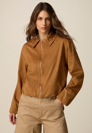 Giovane donna con lunghi capelli castani che indossa una giacca marrone con zip e pantaloni beige, posando con una mano in tasca su uno sfondo uniforme.