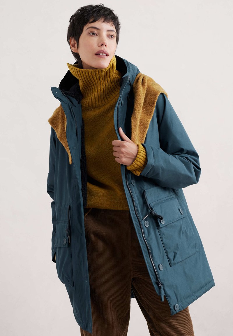 Grove waterproof jack met een rits en knoopsluiting, twee voorzakken; gedragen over een mosterdkleurige coltrui en een bruine corduroy broek.