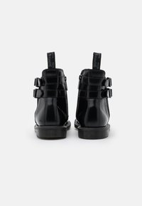 Botas de tobillo de charol negro con dos correas con hebillas a cada lado, una suela de goma texturizada y lengüetas para facilitar su colocación.