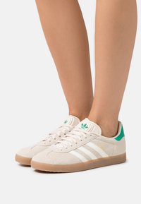 Ben som bär beige Adidas Gazelle-sneakers med vita ränder, gummisulor och gröna hälflikar mot en vit bakgrund.