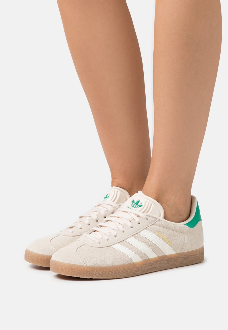 Ben som bär beige Adidas Gazelle-sneakers med vita ränder, gummisulor och gröna hälflikar mot en vit bakgrund.