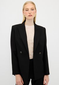 Blazer doppiopetto nero con rever affilati, dettagli in metallo dorato e spalle strutturate, indossato sopra un top in pizzo chiaro.