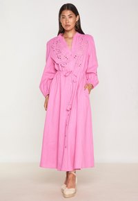 Robe chemise rose avec de longues manches bouffantes, détail de broderie sur le buste, taille à cordon de serrage et jupe longueur cheville. Espadrilles plates portées.