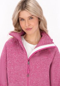 Giacca a zip rosa con finitura testurizzata, dotata di un colletto isolato e dettagli in piping bianco. Il modello indossa una maglietta bianca sotto.