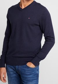 Granatowy sweter v-neck wykonany z miękkiej dzianiny, z małym logo na klatce piersiowej oraz ściągaczami przy mankietach i na dolnym końcu.