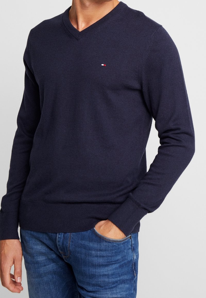 Granatowy sweter v-neck wykonany z miękkiej dzianiny, z małym logo na klatce piersiowej oraz ściągaczami przy mankietach i na dolnym końcu.