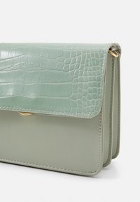 Zelená kabelka z umělé kůže s krokodýlím vzorem na klopě, zlatými kovovými akcenty a strukturovaným tvarem. Minimalistický a elegantní design.