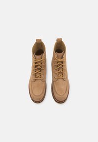 Botas de tobillo de ante color beige con una textura suave, punta redonda y cordones marrones. Presentan un forro de tela y una pestaña de tira en la parte trasera.
