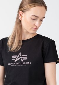 Juodas medvilnės marškinėlis su atspindinčiu logotipu, juostelėmis ir sidabriniais užrašais "ALPHA INDUSTRIES". Trumpomis rankovėmis, klasikinio silueto.