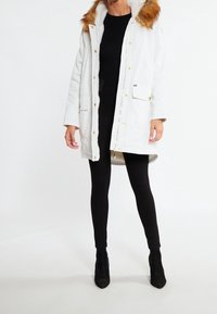 Biała kurtka parka z kapturem wykończonym futerkiem, zamkiem błyskawicznym i zatrzaskami, dwoma przednimi kieszeniami oraz podszytym wnętrzem. Noszona na czarnych legginsach i obcasach.