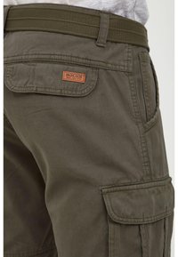 Close-up of olive green cargo shorts with flap pockets and a woven belt, showing a small brown brand patch on the back pocket.
