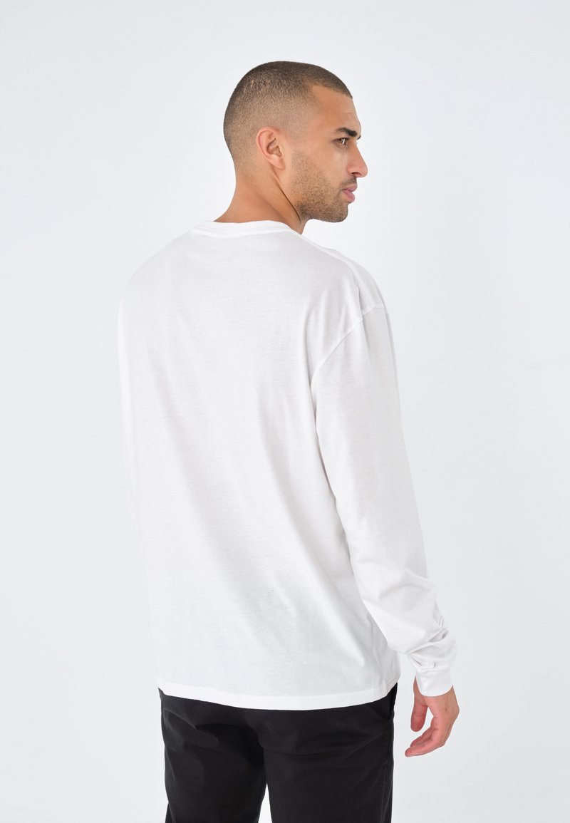 Camisa blanca de manga larga hecha de una tela suave, con un corte relajado y escote redondo. La vista trasera muestra un diseño sin costuras y puños acanalados.