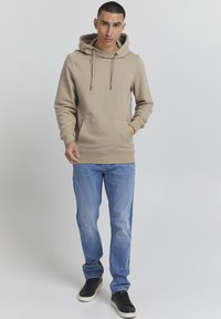 Bež hoodie s podesivim vezicama, prednjim džepom, u kombinaciji sa svijetloplavim trapericama i crnim tenisicama. Glatka tkanina i opušteni kroj.