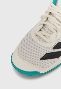 Sneaker with a beige mesh upper, black accents, and a turquoise sole. Features a textured design and traditional lace closure.