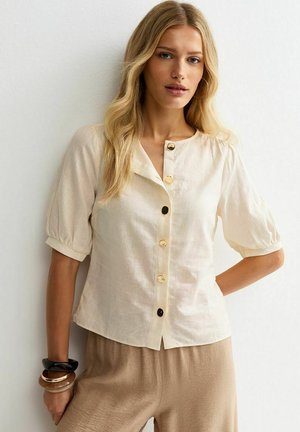 Blondynka w kremowej, krótkiej, zapinanej na guziki bluzce z bufiastymi rękawami i beżowych spodniach, stojąca na tle białej ściany.