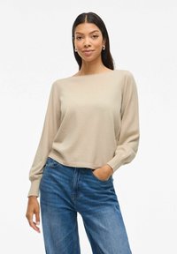 Beige lange mouwen top met een relaxte pasvorm en ronde halslijn, gecombineerd met blauwe high-waisted jeans. De stof lijkt zacht en glad.