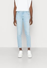 Jeans slim fit azul claro com bainhas dobradas, confeccionados em denim de textura suave. Combinados com uma blusa branca sem mangas e ténis brancos.