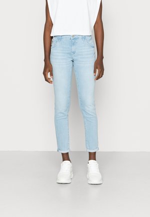 Jeans slim fit azul claro com bainhas dobradas, confeccionados em denim de textura suave. Combinados com uma blusa branca sem mangas e ténis brancos.
