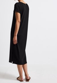 Kobieta w czarnej, prążkowanej sukience midi z krótkimi rękawami oraz bocznym rozcięciem, w połączeniu z brązowymi sandałami ze kwadratowym noskiem, stojąca na białym tle.