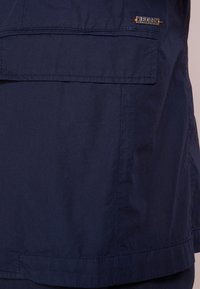 Granatowy płaszcz z gładką fakturą, z klapkową kieszenią i małym akcentem logo. Prosty design z czystymi liniami i szwami.