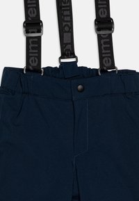 Mørkeblå bukser med sorte justerbare seler. Glatt stoff, elastisk midje og en frontlomme med trykknapplås.