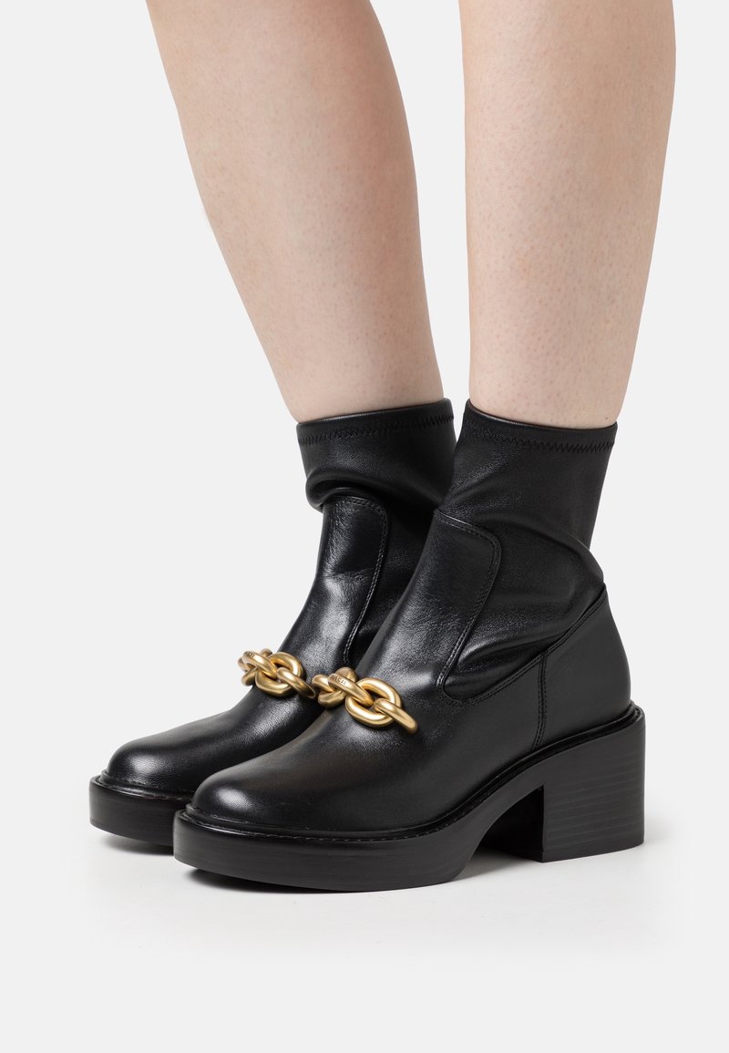 Bottines en cuir noir à talons blocs avec une grande chaîne dorée sur le devant, portées sur jambes nues sur fond blanc.