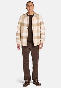 Beige en tan geruite overhemdjas met knopen aan de voorkant, gedragen over een donkerbruine trui, gecombineerd met bruine ribfluwelen broek en bruine schoenen.