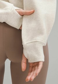 Světle béžová mikina s žebrovanými manžetami, v kombinaci s hnědými legíny. Zaměření na texturu a design rukávů; ruka odpočívající na látce.