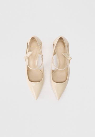 Μπεζ παπούτσια ψηλοτάκουνα από λουστρίνι με μυτερές μύτες, διασταυρωμένες ιμάντες στον αστράγαλο και λεπτομέρεια φιόγκου σε κάθε ιμάντα. Λείος υφής.
