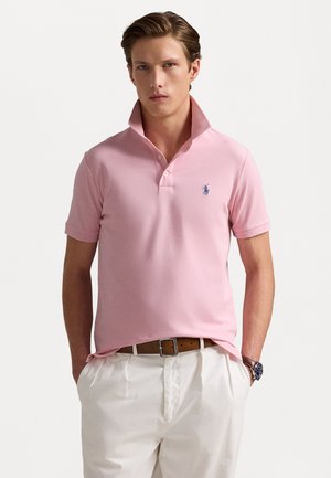 Cămașă polo roz deschis cu un guler lejer, mânecă scurtă și logo brodat pe pieptul stâng, asortată cu pantaloni bej deschis.