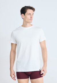 Homme à la peau claire et aux cheveux bruns portant une chemise blanche à manches courtes et un short rouge bordeaux, se tenant devant un fond blanc.