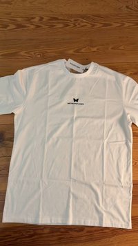 Camiseta de algodón blanca con un logo de mariposa negra en el pecho. Mangas cortas y un corte relajado, colocada plana sobre una superficie de madera.