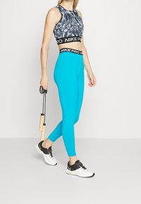 Reggiseno sportivo Nike Pro grigio con motivo astratto e leggings turchesi a vita alta; scarpe da ginnastica bianche e nere; in mano una corda per saltare.